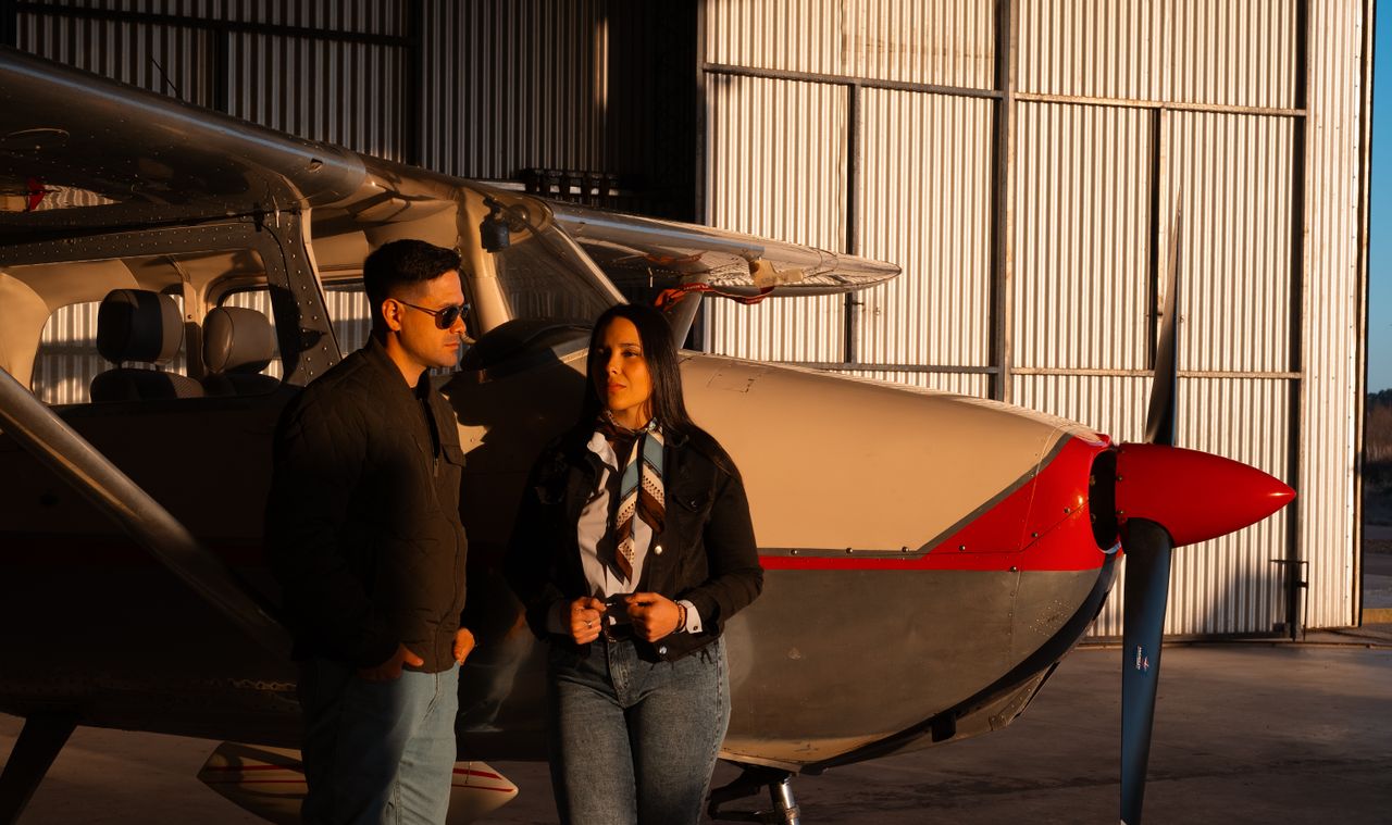 A fashionable couple standing by a small aircraft in a sunlit hangar.
